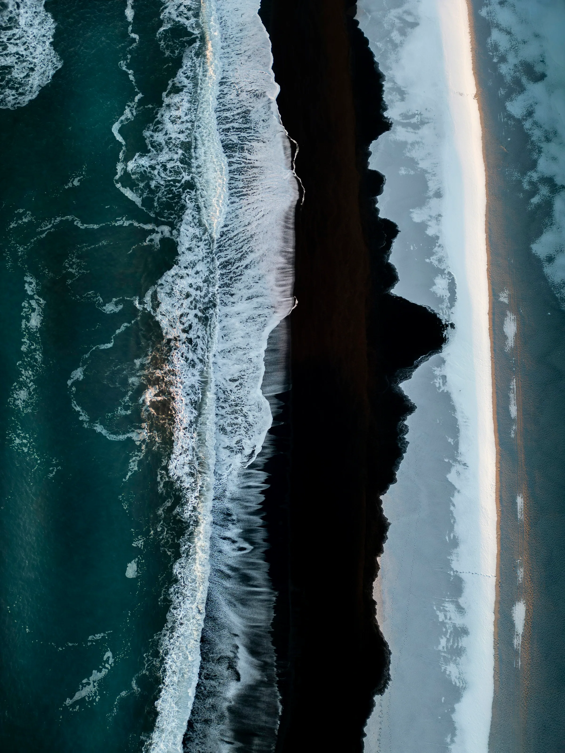 Aerial view of pale surf breaking in long lines across a dark, wet coastline