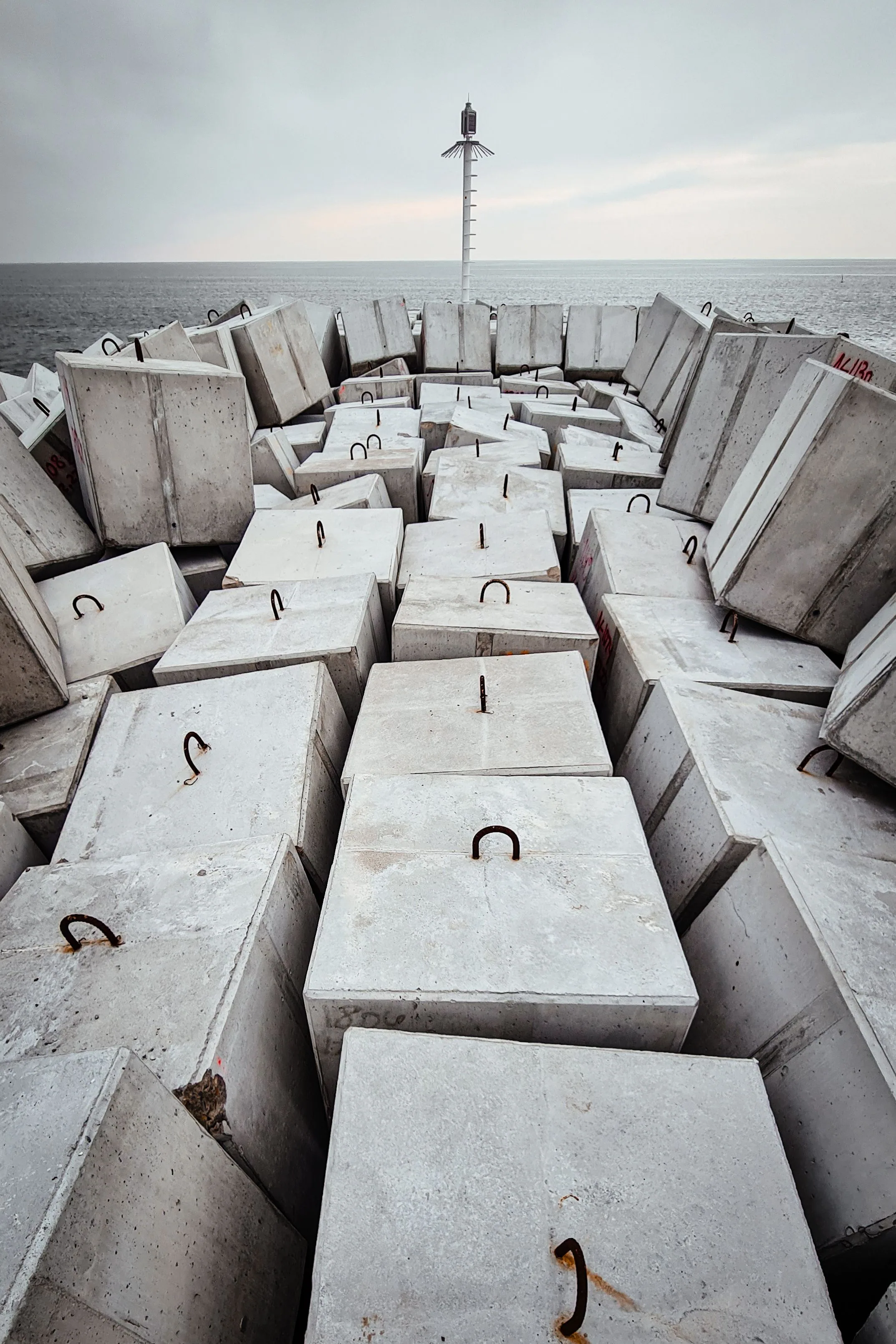 Cast concrete blocks stacked along a shoreline under an overcast sea, a navigation beacon on the horizon